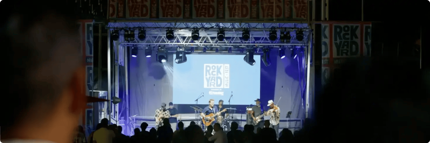 A small band performs at the RockYard Music Fest stage under blue and purple lights, with a bright logo screen behind them and audience silhouettes in the foreground. A small band performs at the RockYard Music Fest stage under blue and purple lights, with a bright logo screen behind them and audience silhouettes in the foreground.